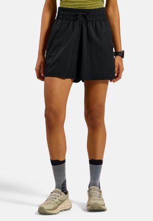 Persoon die zwarte atletische shorts met elastische taille, grijs gestreepte sokken, beige hardloopschoenen en een groen topje draagt, staand op een witte achtergrond.