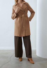Manteau marron sur mesure avec une taille ceinturée, des manches longues et un double revers. Associé à un pantalon large marron et des bottines marron à talons.