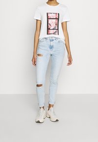 Camiseta blanca con diseño de estampado floral, combinada con unos pantalones vaqueros ajustados rotos de color azul claro y zapatillas beige; atuendo casual acentuado por texturas y colores.