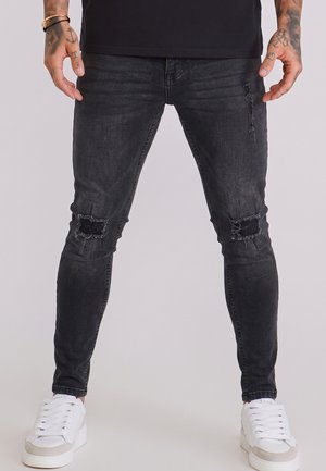 Man draagt zwarte gescheurde skinny jeans en witte sneakers, staat met ontspannen handen langs zijn lichaam tegen een effen achtergrond.