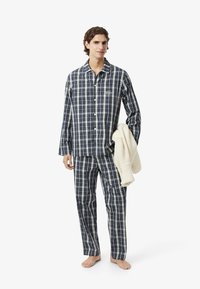 Pigiama da uomo a quadri in blu navy, verde e bianco. Include camicia con bottoni e pantaloni abbinati. Il modello tiene in mano una coperta in maglia color crema.