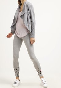 Šedá strukturovaná bunda s přehnutým límcem, doplněná světle šedými legínami s květinovým vzorem na manžetách a bílými teniskami.