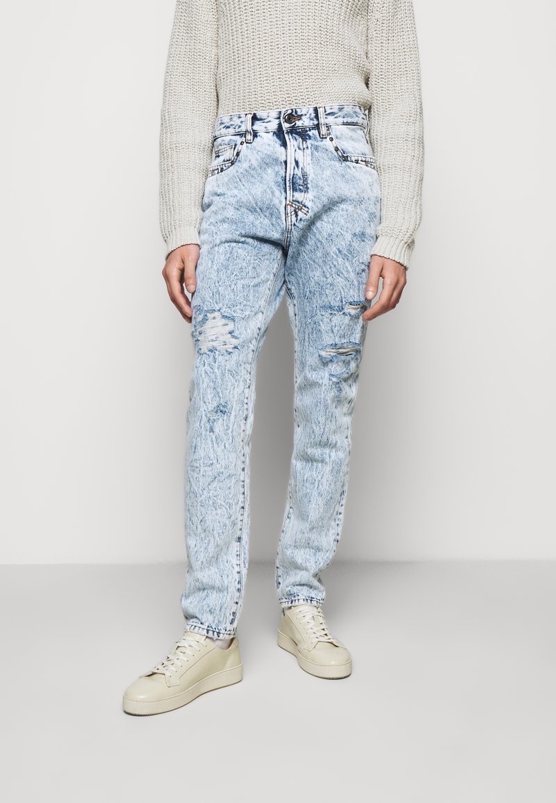 Jeans de mezclilla azul claro con ajuste ajustado, detalles desgastados, textura descolorida, cinco bolsillos y cierre de botón. Combinados con zapatillas beige.