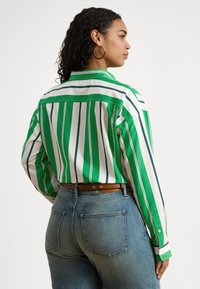 Camisola às riscas verdes e brancas com mangas compridas, vista de costas. Apresenta gola e punhos com botões, combinada com jeans azul-claro e um cinto castanho.