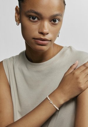 Pulsera de cuentas con perlas blancas redondas, con un cierre de toggle de plata. Llevada en una muñeca delgada contra una blusa de color beige claro.