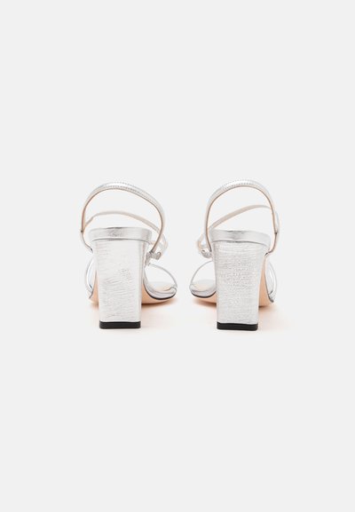 Paire de sandales argentées à talons hauts avec brides à la cheville, vues de dos sur fond blanc.