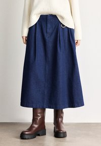 Donkere denim maxi-rok met plooien en een gouden ringdetail op de tailleband. Gecombineerd met bruine enkellaarsjes met dikke zolen.