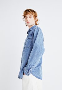 Cămașă din denim de culoare albastră deschisă, cu un buzunar frontal, guler cu nasturi și mâneci lungi. Textură moale cu detalii de cusătură vizibile.