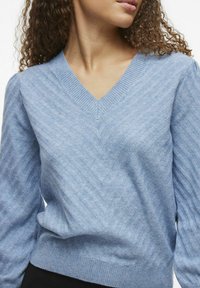 Helles blaues Strickpullover mit V-Ausschnitt, der ein strukturiertes Zickzackmuster sowie gerippte Bündchen und Saum aufweist.