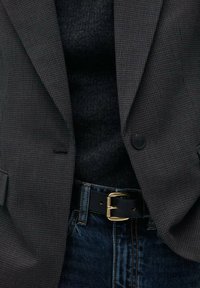 Grå hundtandsmönstrad kavaj med slag, två knappar och sidofickor, bärs över en svart turtleneck och blå jeans med ett bälte med gul spänne.