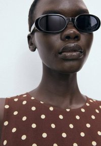 Black oval sunglasses with a thick frame; worn with a brown sleeveless dress featuring cream polka dots. Smooth skin texture visible.