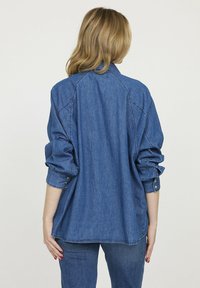 Chemise en denim, coupe décontractée, couleur bleue, manches longues retroussées jusqu'au coude, détails de boutons aux poignets, vue arrière montrant la couture et le design du col.