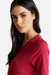 Młoda kobieta z długimi ciemnymi włosami, ubrana w czerwony top z długim rękawem, zwrócona lekko w lewo na tle jednolitego białego tła.