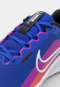 Modré sportovní boty s texturovaným síťovaným svrškem, růžovými akcenty, černými tkaničkami a bílým logem swoosh s barevným mezipodrážkou.