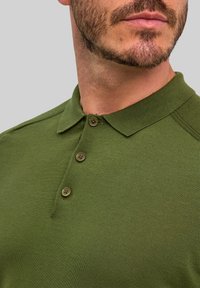 Polo w oliwkowej zieleni z teksturowanej tkaniny, z trzema brązowymi guzikami i klasycznym kołnierzykiem. Zdobione szwem na ramionach dla dodatkowego efektu.