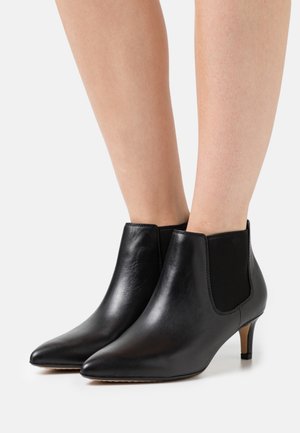 Sorte læderankelstøvler med spids tå og små hæle, båret på bare ben mod en hvid baggrund.