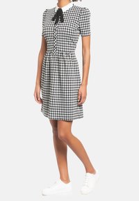 Mekko, jossa on musta-valkoinen ruudullinen kuosi, lyhyet hihat, kaulukset ja nappisuljenta edessä sekä solmimisnauha kaulassa. Yhdistetty valkoisiin tennareihin.