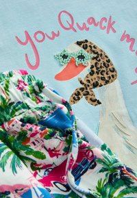 Gęś ubrana w szalik w panterkę i okulary przeciwsłoneczne w kształcie gwiazdy ozdobione koralikami, z różowym napisem „You Quack me” na jasnoniebieskiej tkaninie obok kolorowej tropikalnej tkaniny.