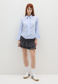 Camicia a righe blu chiaro con colletto e cravatta, abbinata a una gonna nera a righe con volant. Calze bianche e sneaker completano l'outfit.