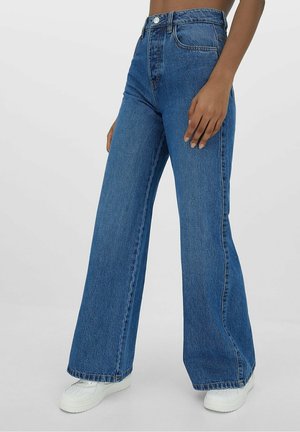 Personne portant un jean bleu taille haute à jambes larges et des baskets blanches, debout devant un fond clair uni.