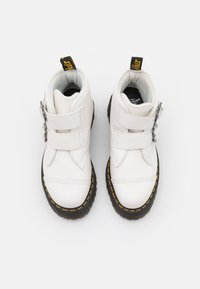 Bottes de cheville en cuir blanc avec deux bandes Velcro, semelle en caoutchouc noir, coutures jaunes et doublure noire. Comprend un accent métallique décoratif.
