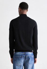 Man with short curly hair wearing a black long-sleeve sweater and blue faded jeans, facing away against a plain white background.