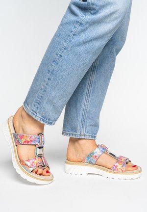 Füße in bunten Sandalen mit floralen Mustern und doppelten Schnallenriemen, kombiniert mit hellblauen, gekürzten Jeans auf weißem Hintergrund.