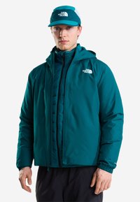 Jeune homme portant une veste The North Face couleur sarcelle et une casquette assortie sarcelle et blanche, debout devant un fond uni.