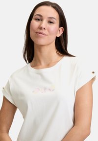 Lys beige bomulds t-shirt med korte rullede ærmer, prydet med et muslingemotiv i pastelfarver på forsiden og guld detaljer med knapper på skuldrene.