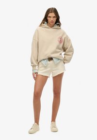 Beżowy, oversize'owy hoodie z logo z przodu, założony na jasnoniebieską koszulę w biało-niebieskie paski, w połączeniu z białymi szortami dżinsowymi i kremowymi sneakersami.