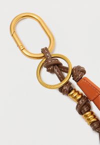 Brūna ādas siksna ar zelta piederumiem, cilpām un mezgliem. Iekļauj oranžu akcentu. Apaļais gredzens ir ar iegravētu zīmolu nosaukumu.