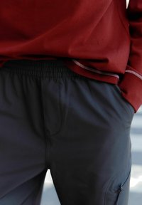 Bordeauxrotes Langarmshirt kombiniert mit dunkelgrauen Hosen mit elastischem Bund, die Seitentaschen und eine Reißverschluss-Tasche am Oberschenkel haben.