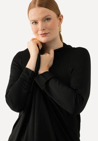 Černý top s dlouhým rukávem vyrobený z hladké tkaniny. Výrazný detail skládání v oblasti výstřihu a přiléhavé manžety. Model drží látku poblíž výstřihu.
