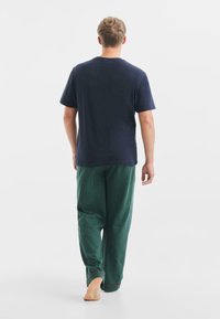 Camiseta de algodón azul marino combinada con pantalones de pijama a cuadros verdes con un acento rojo en el dobladillo. El modelo está descalzo y se ve desde atrás.
