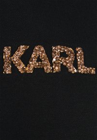 Tela negra con superficie texturizada que presenta el nombre "KARL" en grandes rhinestones dorados. Las letras tienen un aspecto brillante.