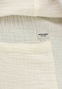 Detailný záber krémovo sfarbené textúrovanej látky s vreckom a vyšitou nášivkou "JACK & JONES EST. 1990".