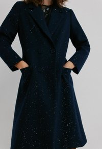 Femme portant un manteau croisé bleu marine foncé avec des détails en sequins, les mains dans les poches, sur un fond clair uni.