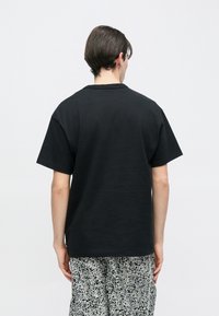 Młoda osoba o ciemnych włosach, ubrana w prostą czarną koszulkę z krótkim rękawem oraz czarno-białe wzorzyste spodnie, stoi tyłem do aparatu.