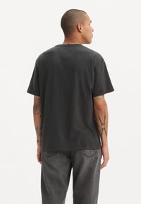 T-shirt en coton gris foncé à manches courtes, col rond et coupe décontractée, associé à un jean gris et présentant des détails de tatouages visibles sur les bras.