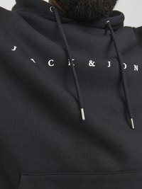 Sort hættejakke lavet af blødt stof med en kengurulomme, prydet med hvidt trykt tekst "JACK & JON" foran og snore med metalspidser.