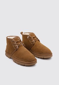Par de botas de ante marrón hasta el tobillo con cordones y forro de piel blanca suave, sobre un fondo gris claro.