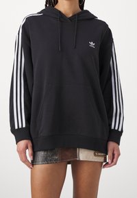 Svart huvtröja med tre vita ränder på varje ärm, framficka med kengurutaske, och en liten vit Adidas-logotyp på bröstet.