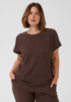 Femme aux cheveux mi-longs portant un haut marron à manches courtes et un pantalon assorti, les mains dans les poches, sur un fond uni.