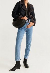 Schwarze Kunstlederjacke mit Pelzkragen, blaue hochtaillierte Jeans und schwarze Stiefeletten. Model posiert mit einer schwarzen Umhängetasche.
