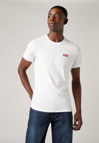Fehér pamut póló rövid ujjakkal, kerek nyakkivágással és egy piros "Levi's" logós sapkával a bal oldali mellkason. Kék farmernadrággal viselve.