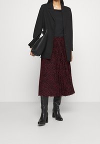 Sort blazer over en sort top, parret med en rød mønstret plisseret nederdel. Sorte hæle støvler og en struktureret sort håndtaske fuldender looket.