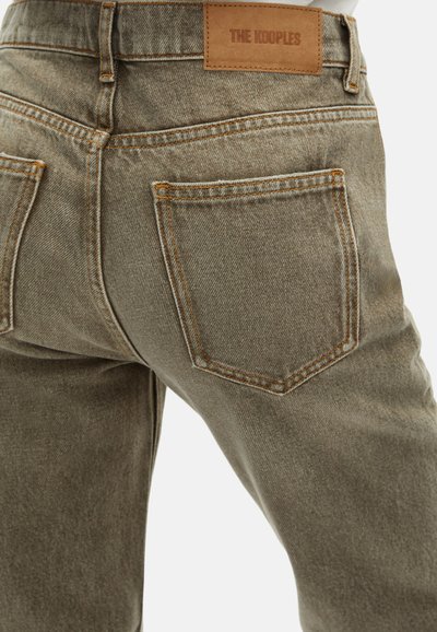 Jeans de mezclilla en un color verde oliva apagado con un acabado texturado, que presentan una etiqueta de marca de cuero y costuras naranjas en contraste.