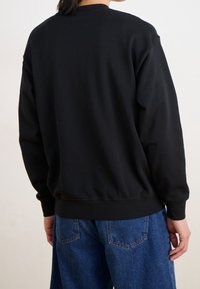 Zwarte sweatshirt met een ronde hals, lange mouwen en geribbelde zoom, weergegeven vanaf de achterkant. Gecombineerd met blauwe spijkerbroek. Eenvoudig, effen kleur.