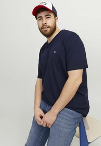 Navy blue short-sleeve t-shirt with a small white logo on the chest, paired with light blue jeans. Model sits on a chair, hands clasped.