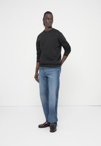 Juodas džemperis su apvalia iškirpte ir ribinėmis rankovėmis, derinamas su šviesiai mėlynomis, tiesiomis kelnėmis bei tamsiai rudos spalvos batais. Paprasta fone.